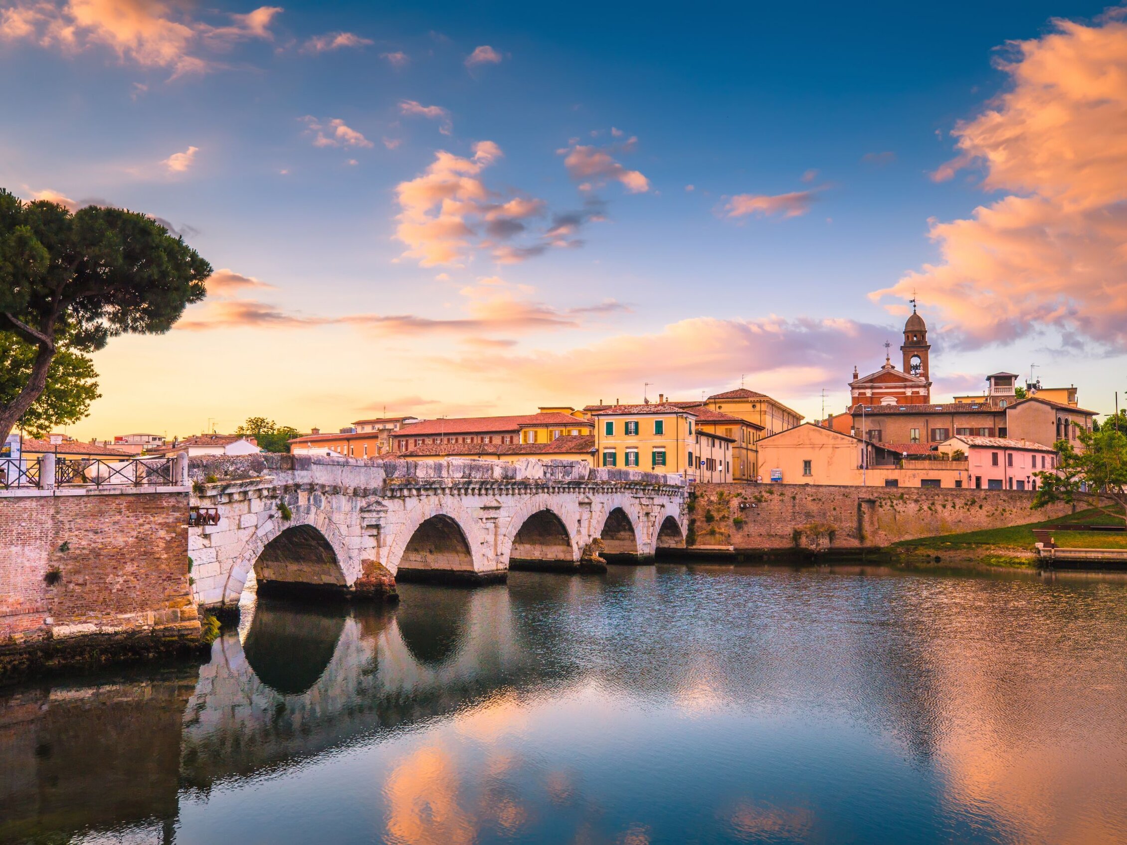 Panorama del Ponte di Tiberio a Rimini al tramonto con riflessi sull’acqua e cielo colorato